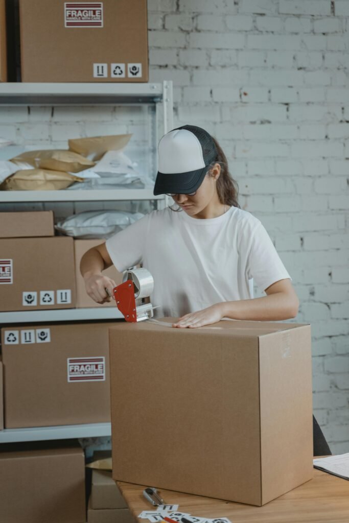 pexels-photo-6169030-6169030 Young female worker sealing cardboard boxes in an indoor warehouse setting.